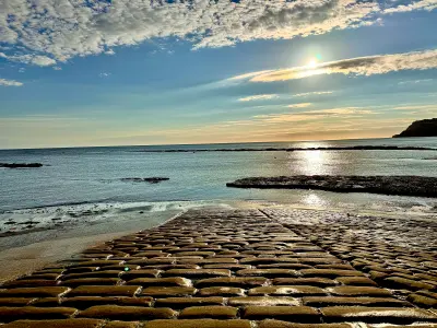 Wet cobblestone slipway leading towards the sea at Robin Hood's Bay, North Yorkshire, with sunlight reflecting across the water beneath a cloud-filled sky.