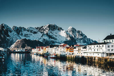 Snow-covered peaks rising behind colourful waterfront buildings and fishing boats in Henningsvær harbour on Austvågøya in the Lofoten Islands, Nordland, Norway.