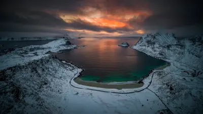 Drone view of Haukland and Vik Beach on Vestvågøya in winter, with snow-covered mountains surrounding a turquoise bay under a dramatic red-orange twilight sky in the Lofoten Islands, Norway.
