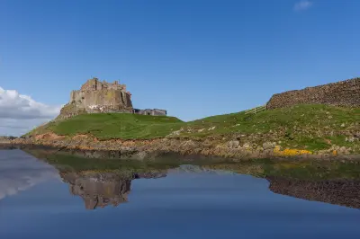 Lindisfarne Castle on a grassy rocky hill overlooking still tidal water, with the castle and surrounding stone walls reflected in the water on Holy Island, Northumberland.