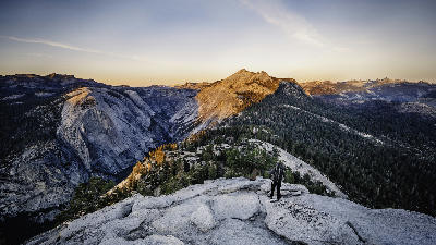 Person standing on a granite overlook above Yosemite Valley with forested ridges and mountains at sunset, California.