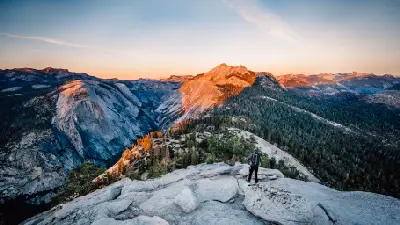 Person standing on a granite overlook above Yosemite Valley with forested ridges and mountains at sunset, California.