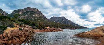 Rugged pink granite shoreline and clear water at Honeymoon Bay with towering granite peaks of The Hazards behind, Freycinet National Park, Tasmania.