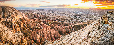 Panoramic sunset view over Red Valley in Cappadocia, Turkey, featuring colourful volcanic cliffs, deep canyons, fairy chimneys, and layered rock formations stretching across the landscape.