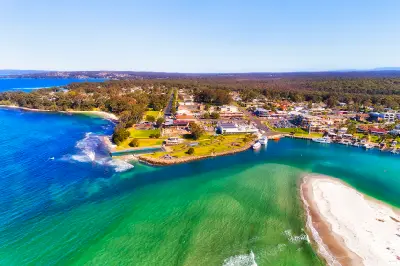 Turquoise waters surround Huskisson’s waterfront and the sandy entrance to Currambene Creek, with the town and forest stretching inland.