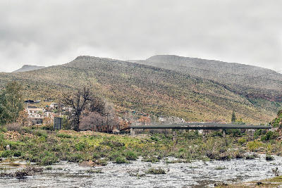 The Tra-Tra River flowing through Wupperthal beneath a pedestrian bridge, with the historic mission settlement and surrounding Cederberg mountains in the background.