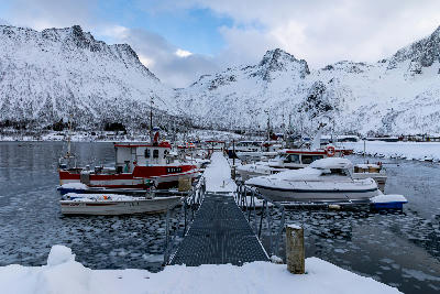 Small winter harbour with fishing boats, icy water, and a pier leading toward snow-covered mountains on Senja island, Norway