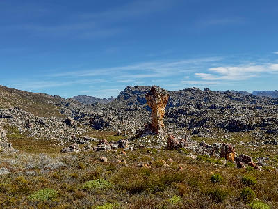 The Maltese Cross rock formation rising from a rugged sandstone landscape in the Cederberg Wilderness Area, surrounded by rocky terrain and mountain ridges under a clear blue sky.