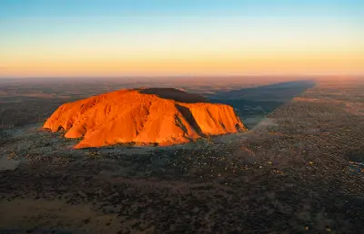 Aerial view of Uluru glowing orange in low evening sunlight, casting a long shadow across the desert in Australia's Northern Territory.