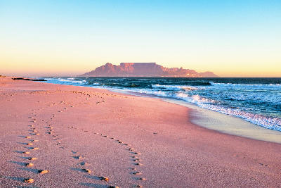 Table Mountain viewed from Bloubergstrand beach at sunset with waves rolling onto the shore and footprints in the sand, Cape Town, South Africa.