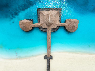 Top-down aerial view of a wooden jetty with traditional thatched-roof structures over turquoise Indian Ocean waters at Nungwi Beach, Zanzibar, Tanzania.