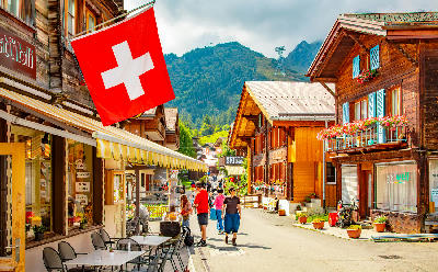 Pedestrian street in Mürren village with wooden chalets, flower balconies, and people walking beneath a Swiss flag, Switzerland.