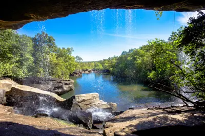 View from behind the curtain of Little Mertens Falls across a tranquil pool surrounded by lush vegetation and sandstone rocks.