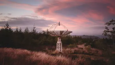 Radio telescope dish surrounded by conifer trees and golden grasses beneath a pink and purple twilight sky at Kielder Observatory, Northumberland.