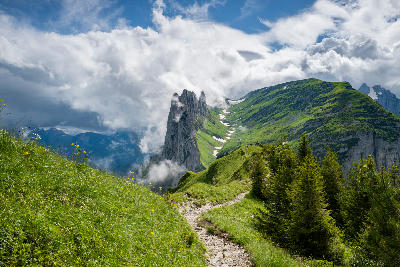 Mountain trail along a grassy ridge leading toward the jagged limestone peaks of Saxer Lücke in the Alpstein mountains, Switzerland, with clouds moving through the scene.
