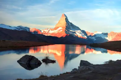 Matterhorn reflected in Stellisee at sunrise with warm alpenglow and foreground rocks, Switzerland.