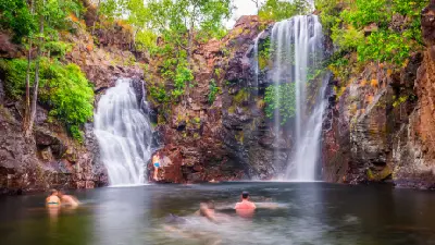Visitors swimming in the natural plunge pool beneath two cascading waterfalls, surrounded by reddish sandstone cliffs and lush green vegetation at Florence Falls, Litchfield National Park, Northern Territory, Australia.