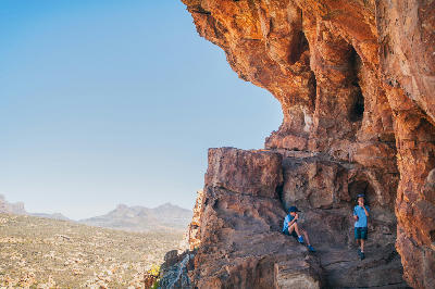 Two young visitors resting beneath a massive sandstone overhang at Truitjieskraal in the Cederberg Wilderness Area, overlooking rugged mountain landscapes and the distant Sneeuberg peaks in the Western Cape, South Africa.