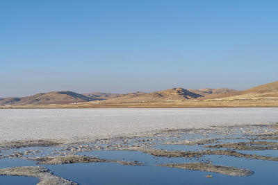 White salt flats and shallow blue pools at Lake Tuz with dry rolling hills in the distance near Şereflikoçhisar, Turkey.