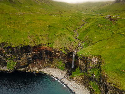 Aerial view of Skarðsáfossur waterfall flowing from a grassy valley onto a rocky beach along the Atlantic coast of Vágar Island, Faroe Islands.