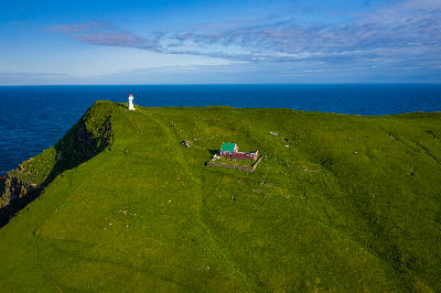 Drone view of Mykines Hólmur Lighthouse and red keeper’s house on a grassy cliff ridge above the Atlantic Ocean, Faroe Islands.