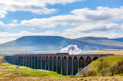 Steam locomotive crossing the stone arches of Ribblehead Viaduct with green hills and blue skies in the Yorkshire Dales, England.