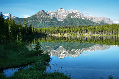 Herbert Lake reflecting forest and layered mountain peaks under clear blue sky in Banff National Park, Alberta, Canada.